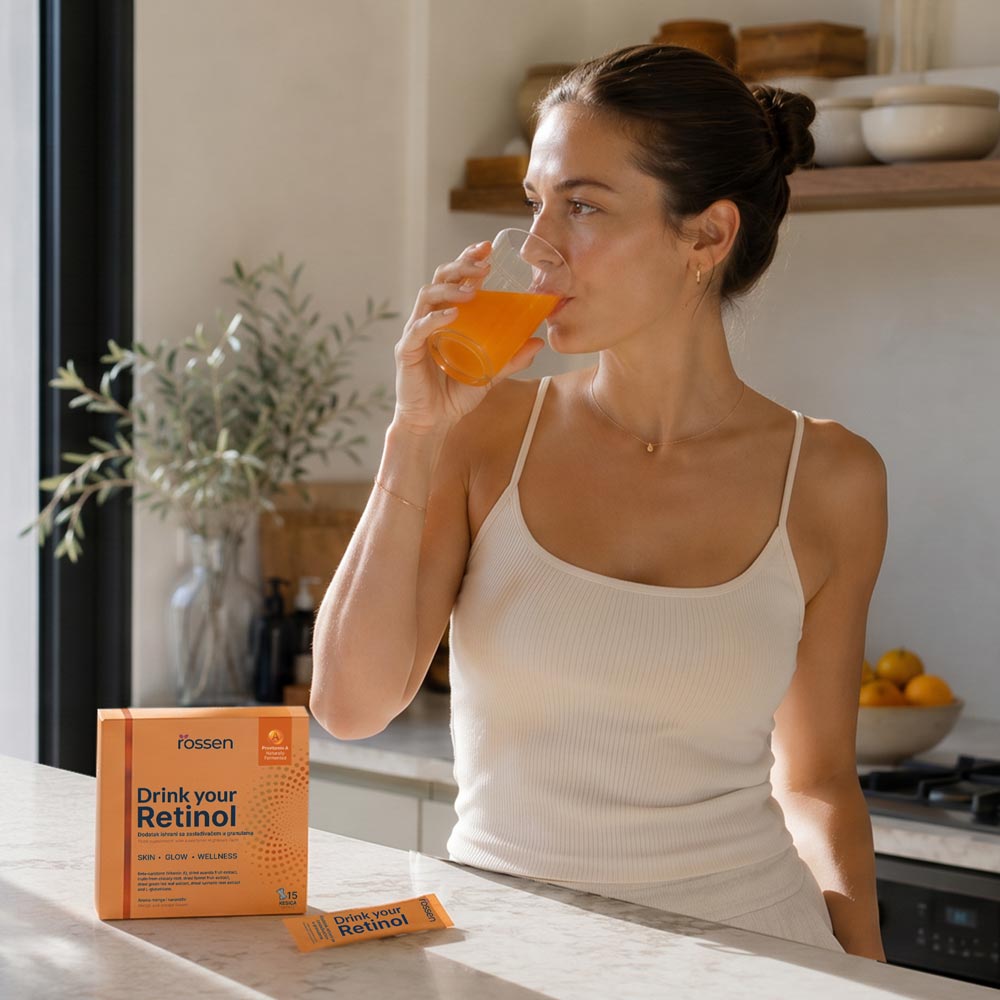 Woman in a white tank top drinks orange juice at a kitchen island; an orange box labeled 'Drink your Retinol' sits on the counter nearby.